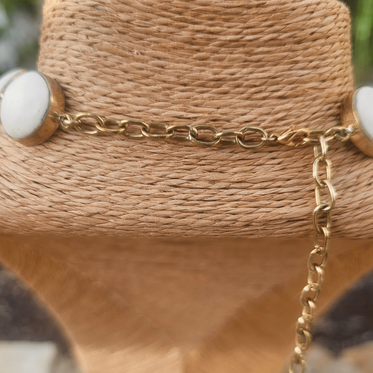 A gold-tone chain necklace with oval white stone accents, displayed on a woven tan mannequin bust with an outdoor blurred background. Shop Kamawe