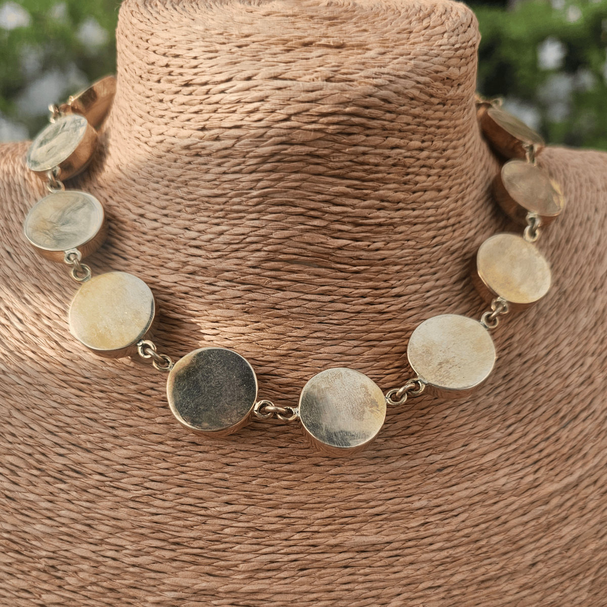 The back side of a statement necklace with large round off-white cabochon stones set in gold-tone metal, displayed on a textured tan woven jewelry bust. Shop Kamawe