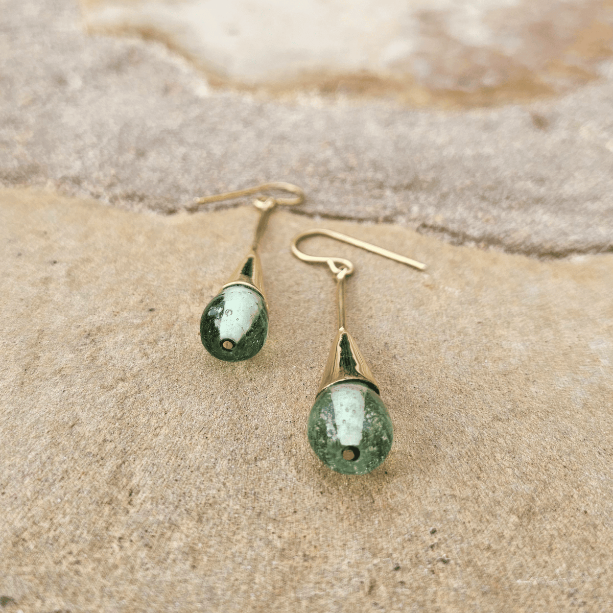 A pair of gold dangle earrings with teardrop-shaped green gemstone drops, resting on a textured beige stone surface. Shop Kamawe