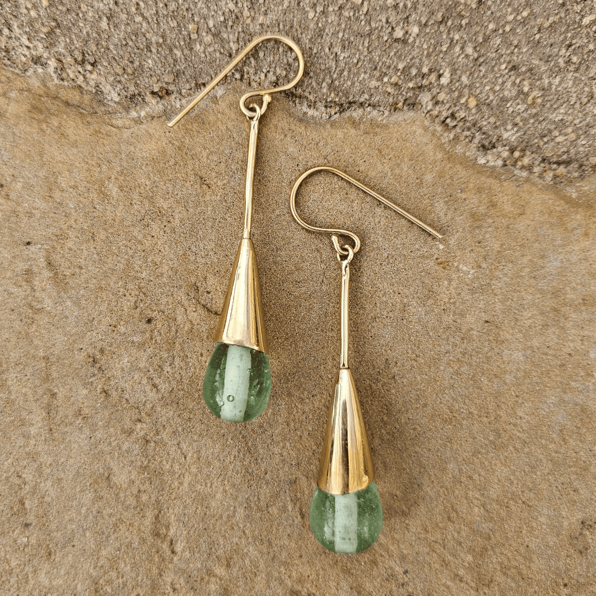 A pair of gold drop earrings with teardrop-shaped green gemstone pendants, displayed on a textured brown stone surface. Shop Kamawe