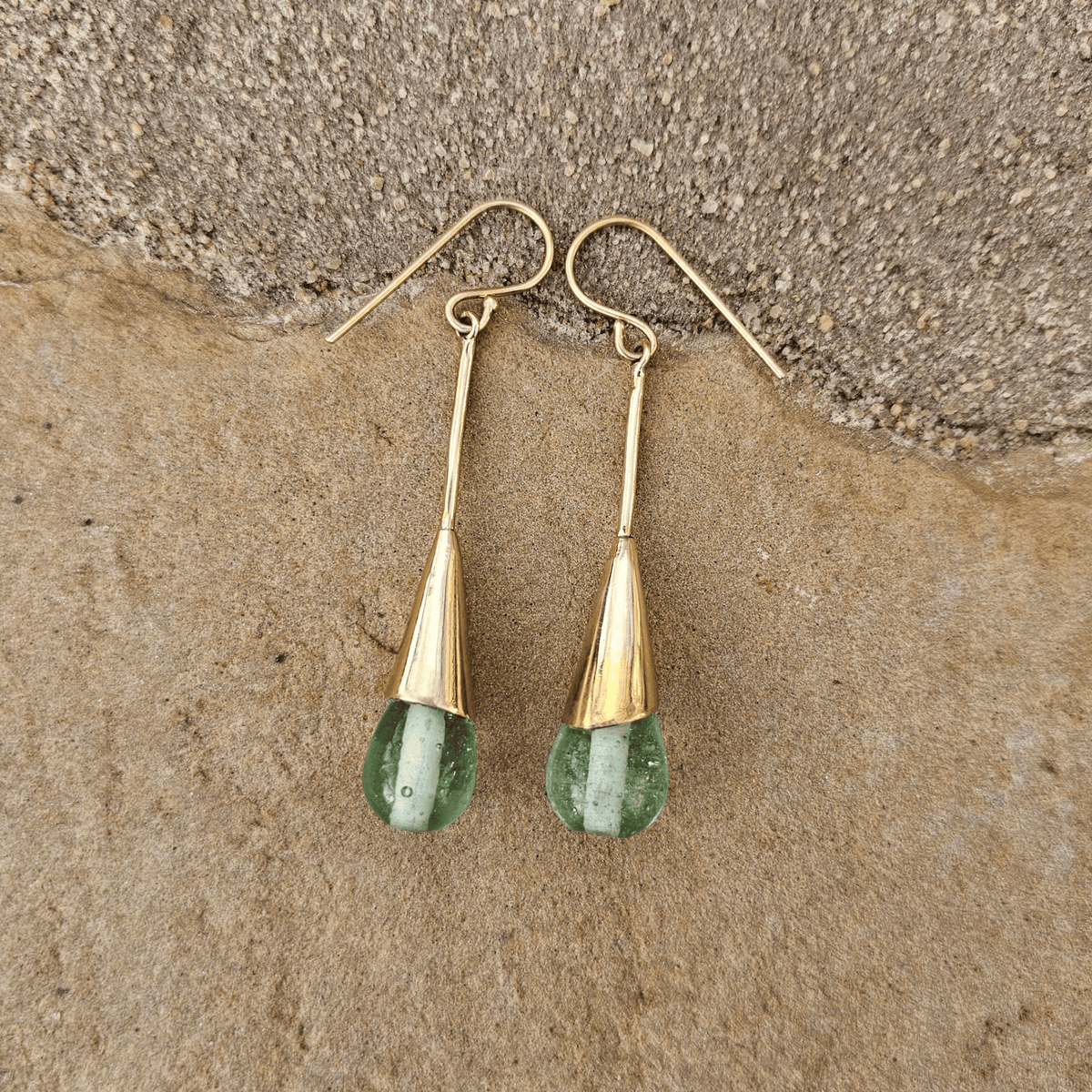 A pair of gold drop earrings with teardrop-shaped green gemstone pendants, resting on a textured brown stone surface. Shop Kamawe