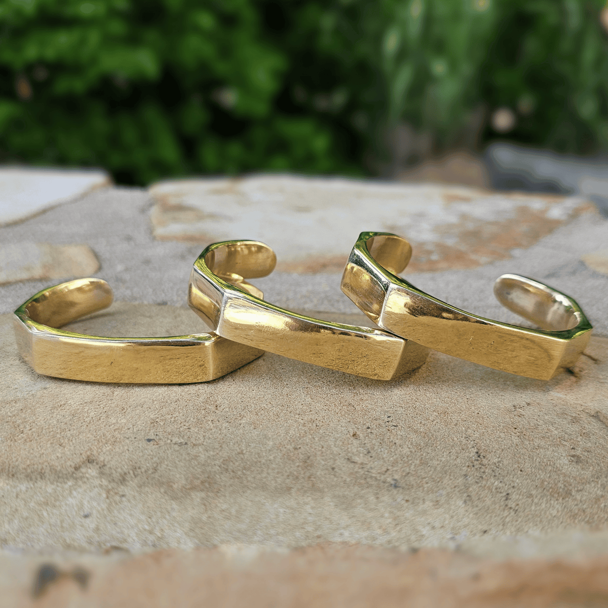 Three gold-toned geometric cuff rings with a textured finish, arranged on a stone surface with a blurred green outdoor background. Shop Kamawe