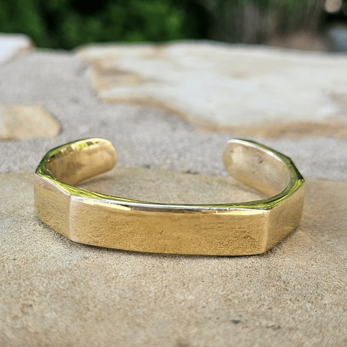 A chunky, open cuff bracelet with a matte gold finish and a geometric, slightly faceted octagonal design, resting on a textured stone surface with a blurred green background. Shop Kamawe