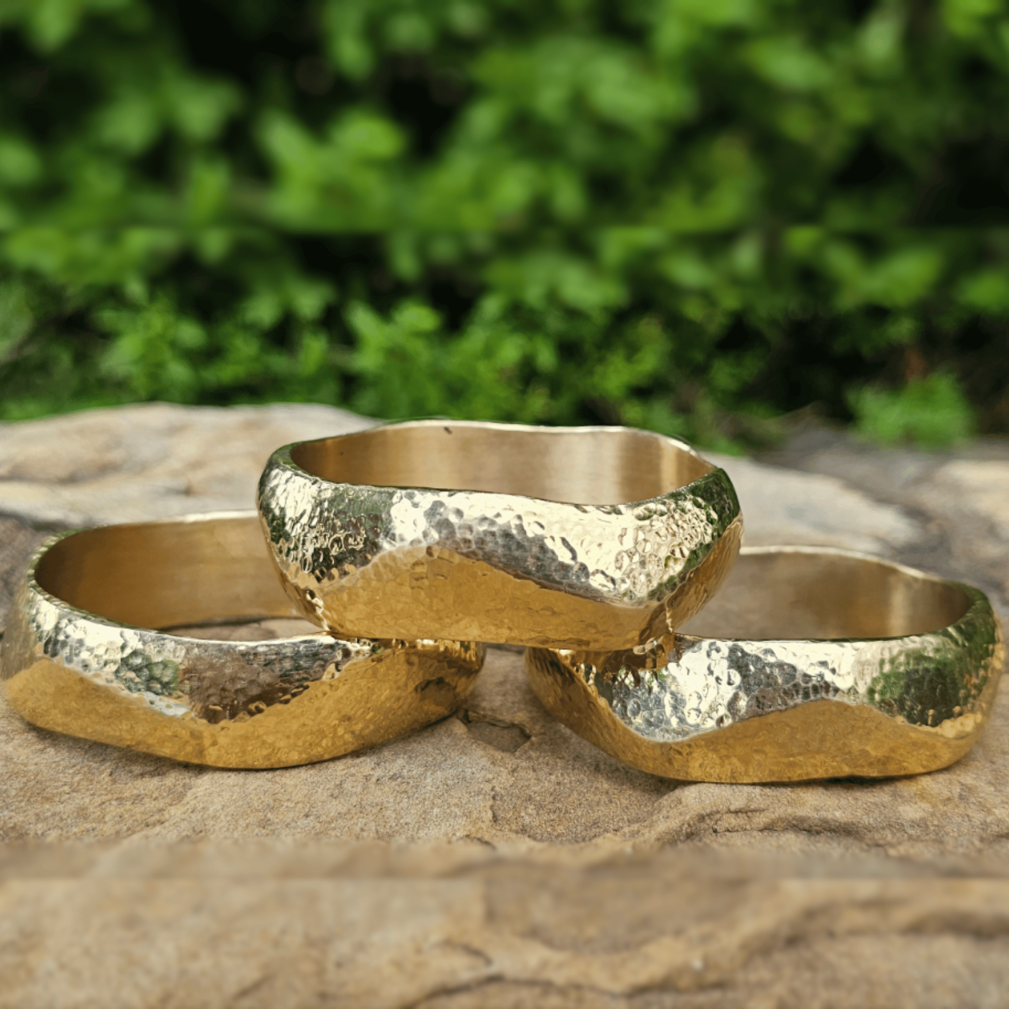 Three hammered gold rings with a wavy, organic shape resting on a stone surface with a blurred green foliage background. Shop kamawe