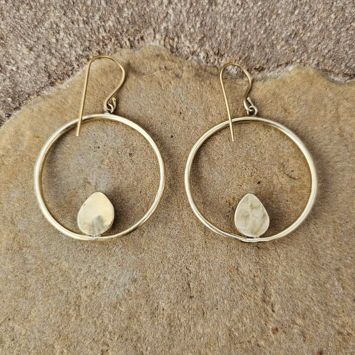 A pair of gold-tone hoop earrings with a small teardrop-shaped stone set in the center of each hoop, displayed on a textured tan stone surface. Shop Kamawe