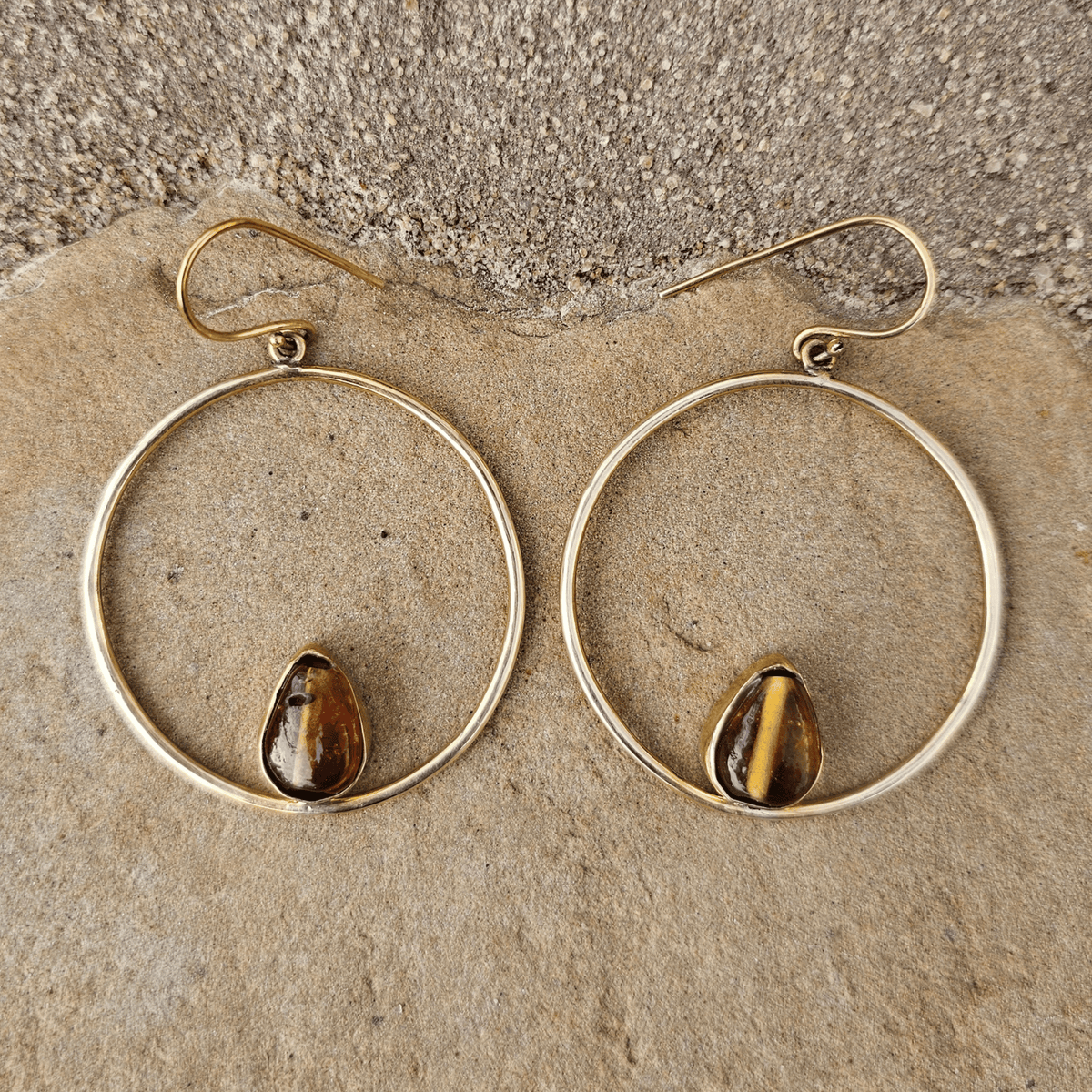 A pair of gold-tone drop earrings with large open circular hoops and teardrop-shaped brown tiger's eye stones, displayed on a textured beige stone surface. Shop Kamawe