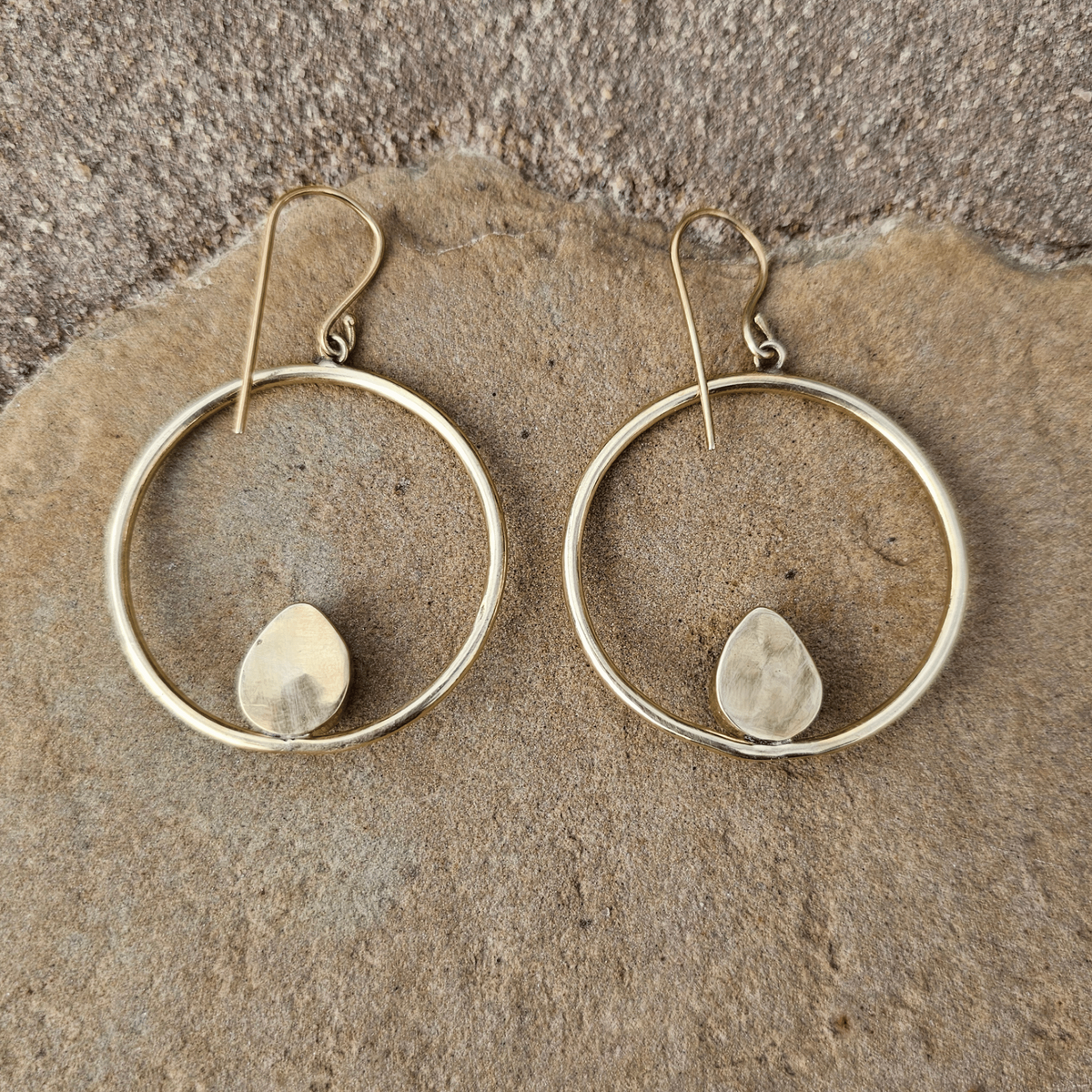 A pair of gold hoop earrings with a small teardrop-shaped purple gemstone set inside each hoop, displayed on a textured beige stone surface. Shop Kamawe