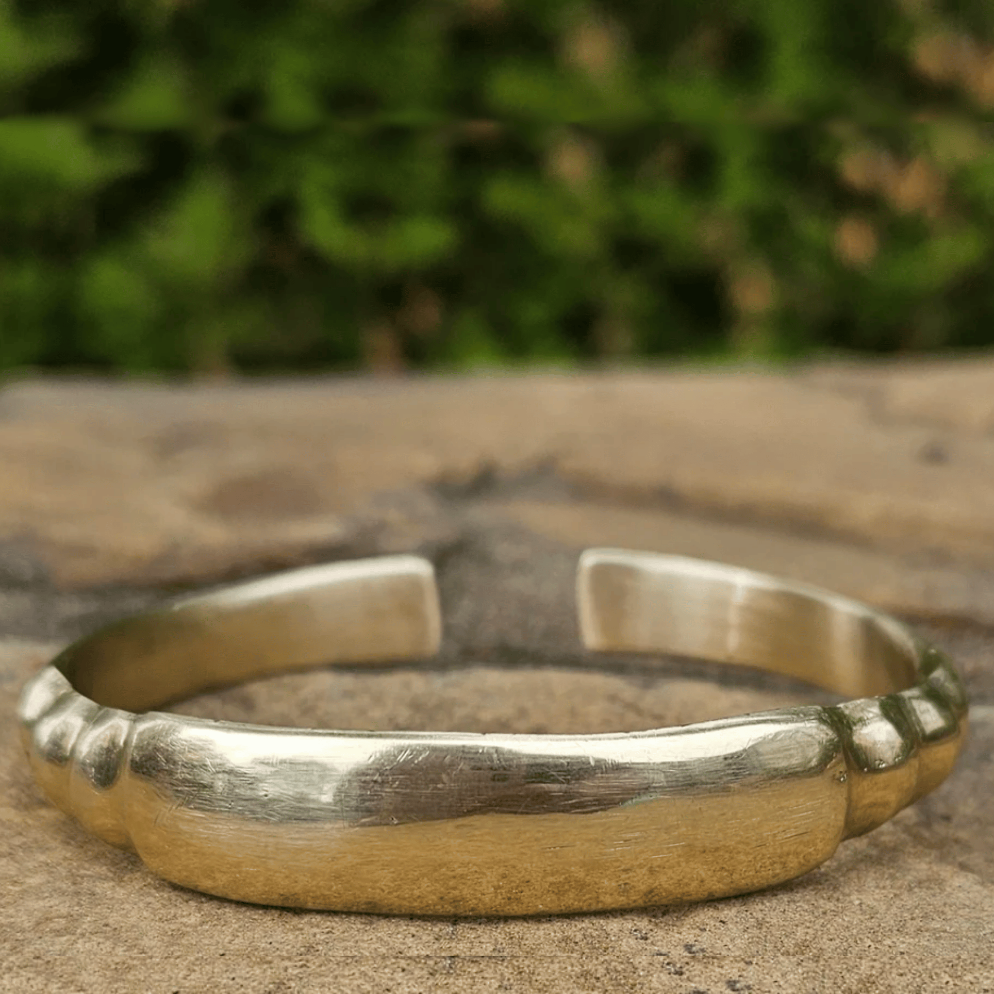 A hammered sterling silver cuff bracelet with a polished finish, resting on a textured stone surface with a blurred green foliage background. Shop kamawe