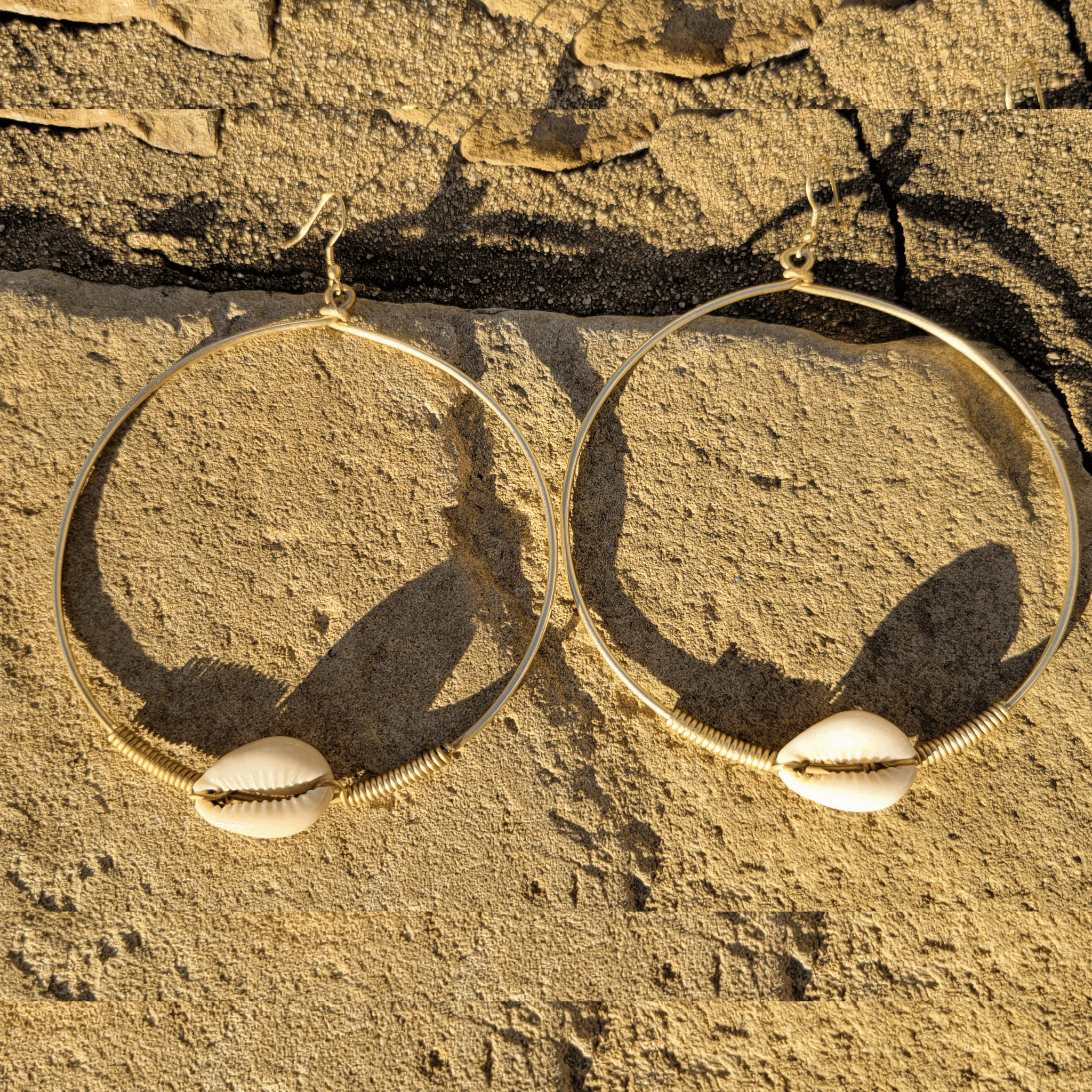 A pair of large gold-tone hoop earrings with cowrie shells at the bottom, displayed on a textured sandy surface. Shop Kamawe