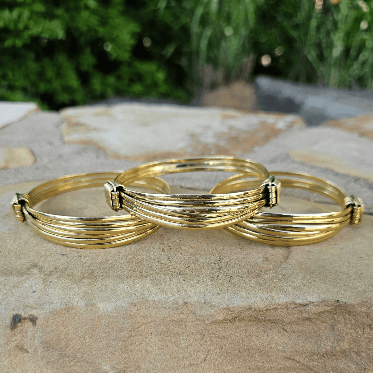 Three gold-toned multi-strand bangle bracelets with small rectangular connector details, arranged on a stone surface with blurred green foliage in the background. Shop Kamawe
