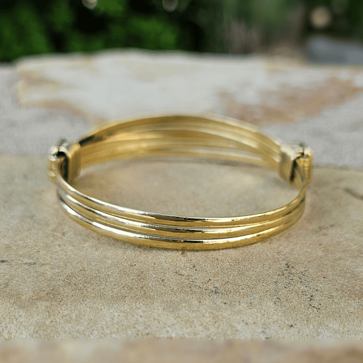 A gold-plated multi-strand bangle bracelet resting on a textured stone surface with a blurred green background. Shop Kamawe