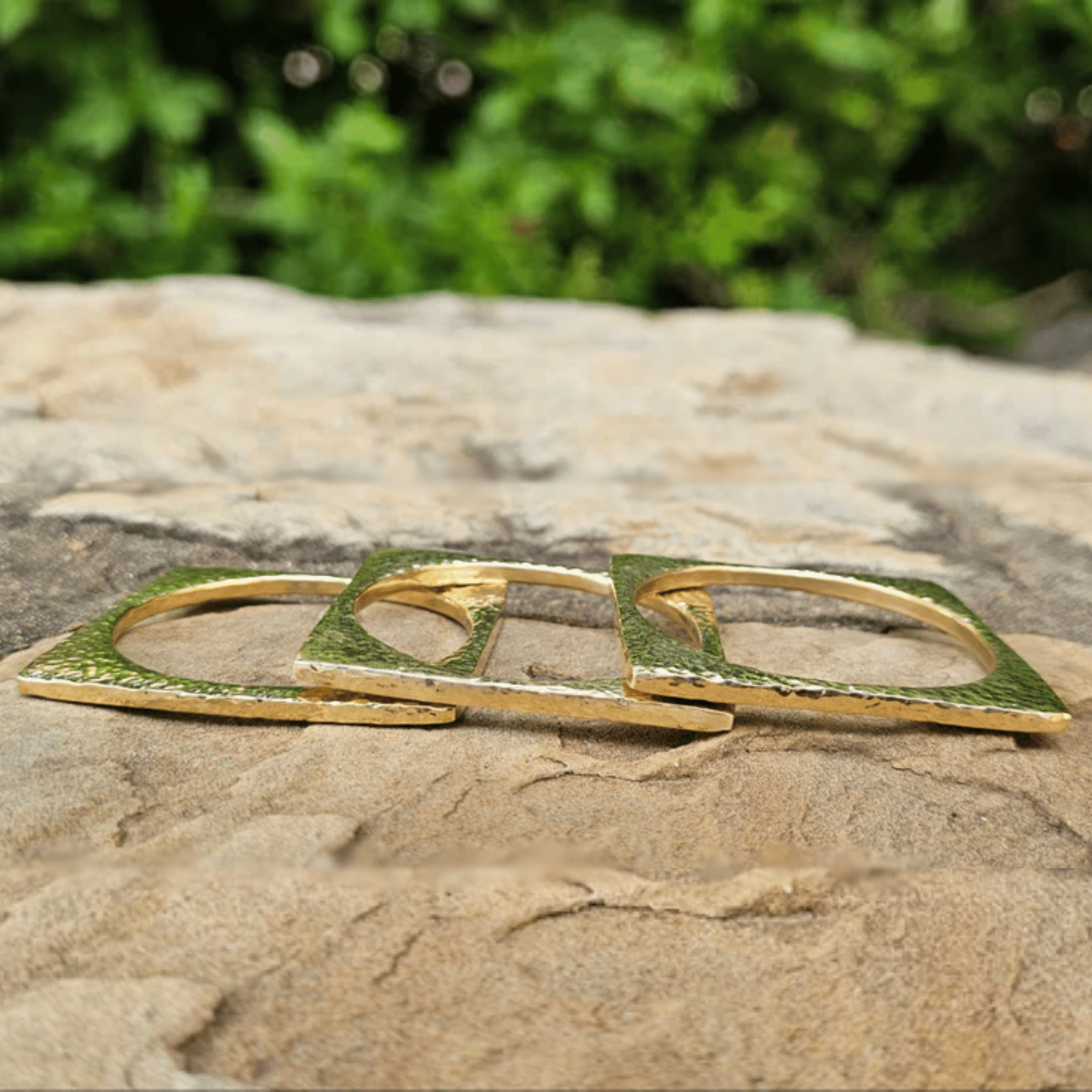 A pair of gold-toned square ring holders or earrings with circular cutouts, resting on a textured stone surface with a blurred green garden background. Shop Kamawe