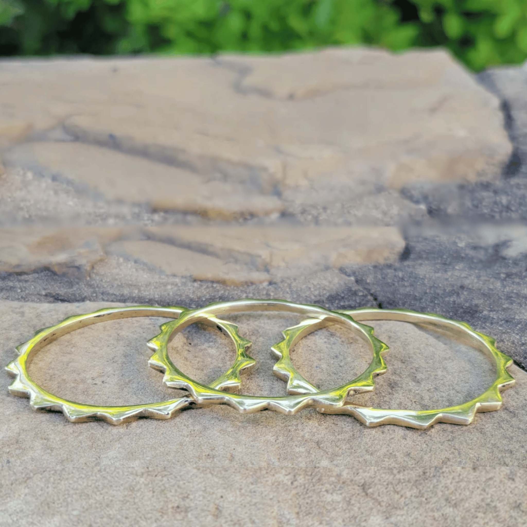 Three gold-toned metal bangles with a jagged, spiked edge design, displayed on a textured stone surface with a blurred green background. Shop Kamawe