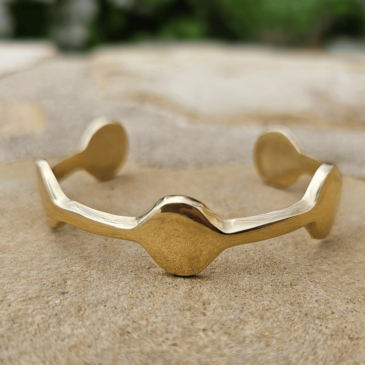 A polished gold cuff bracelet with a wavy, organic shape and rounded ends, resting on a textured beige stone surface. Shop Kamawe