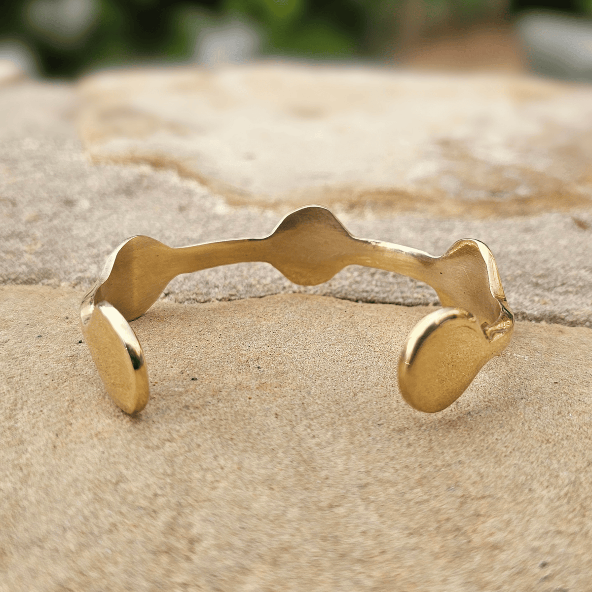 A gold-toned open cuff bracelet with organic wavy edges, resting on a textured beige stone surface. Shop Kamawe