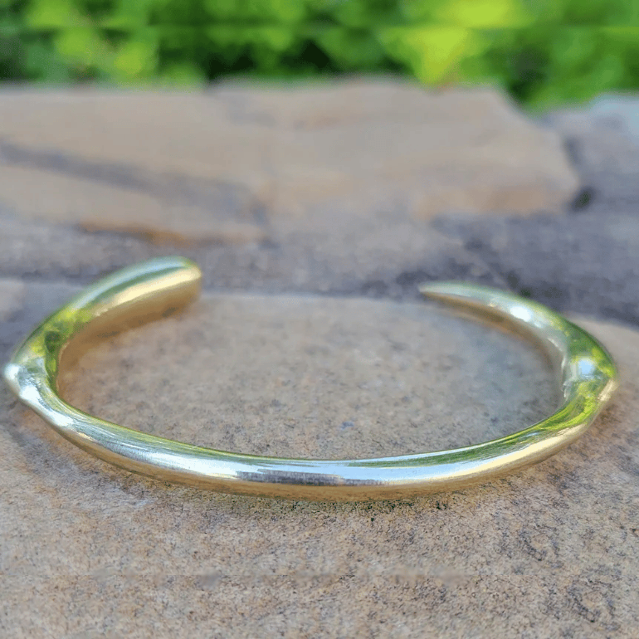 A polished gold-toned metal cuff bracelet resting on a textured stone surface with a blurred green outdoor background. Shop Kamawe