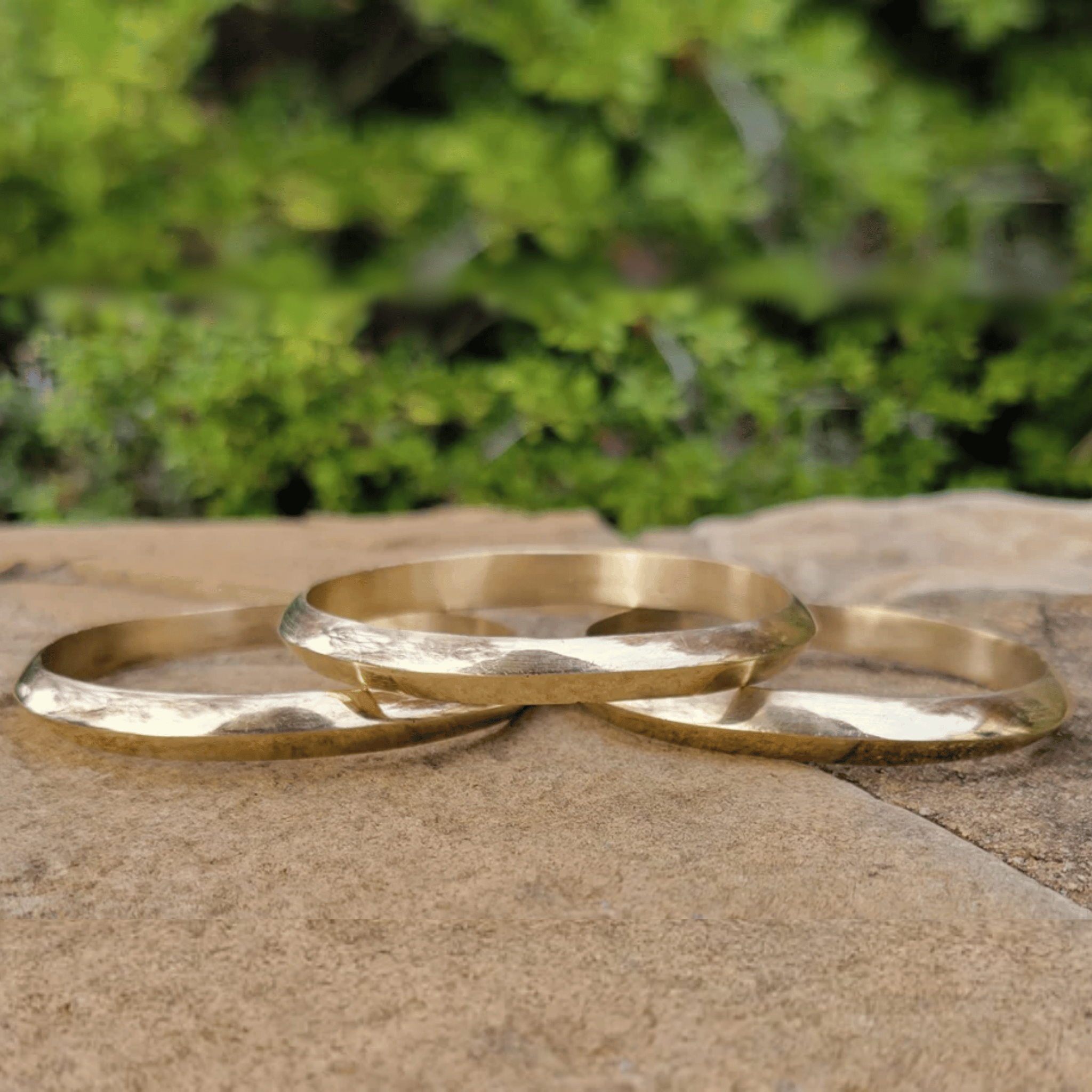 Three hammered gold-tone metal bangles resting on a textured stone surface with a blurred green foliage background. Shop Kamawe