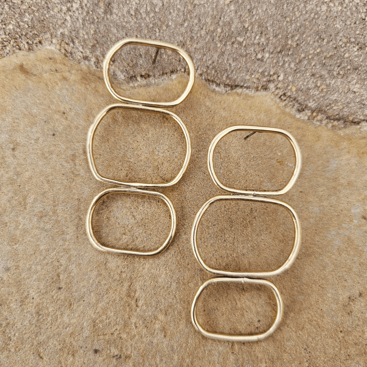 Six gold-toned metal ring blanks with rounded rectangular and square shapes, arranged on a textured tan stone surface. Shop Kamawe
