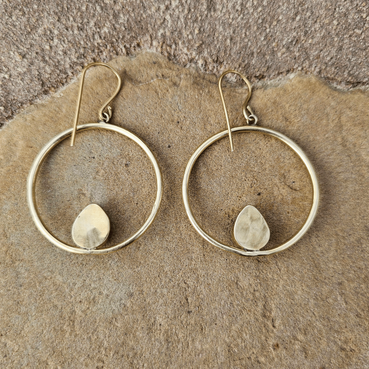 A pair of gold-toned drop earrings with thin circular hoops and small irregular stone pendants, displayed on a textured tan stone surface. Shop Kamawe