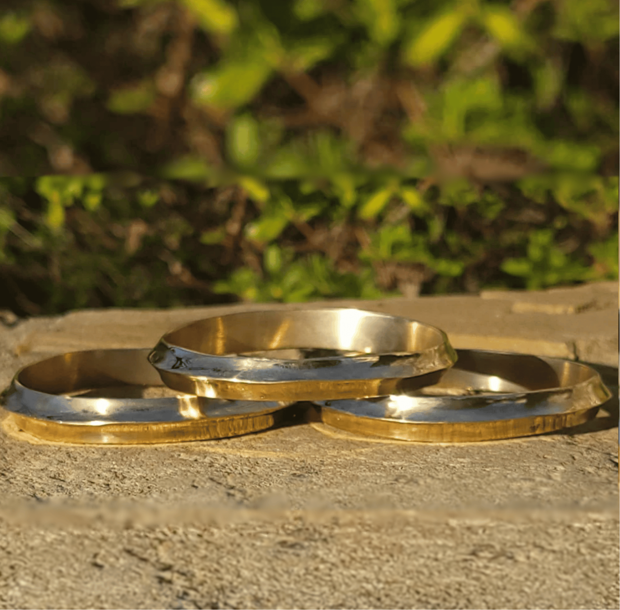 Three metallic bangles with a polished silver center and a textured gold-toned outer rim, arranged on a stone surface with a blurred green outdoor background. Shop Kamawe