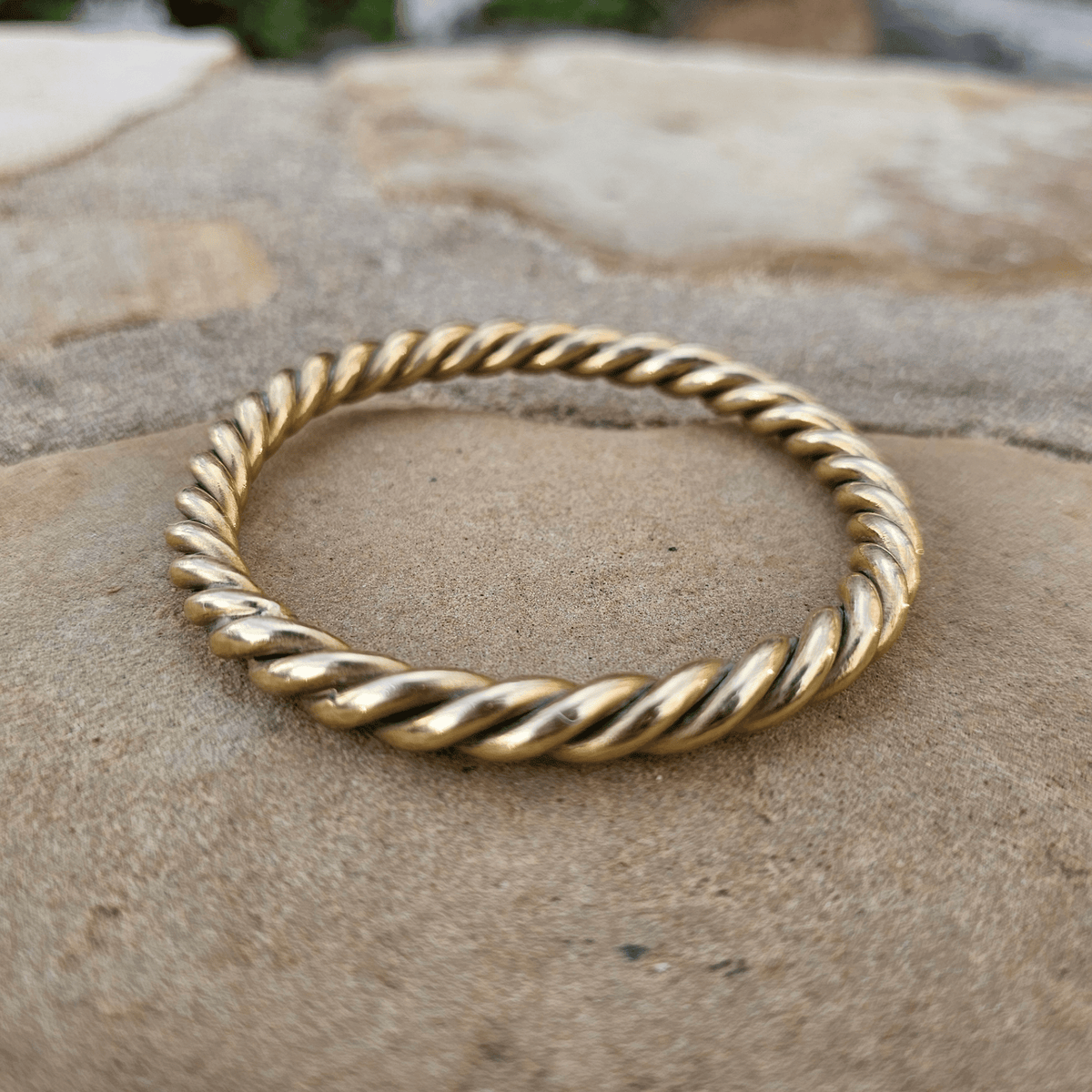 A twisted rope-style gold bangle bracelet resting on a textured stone surface. Shop Kamawe