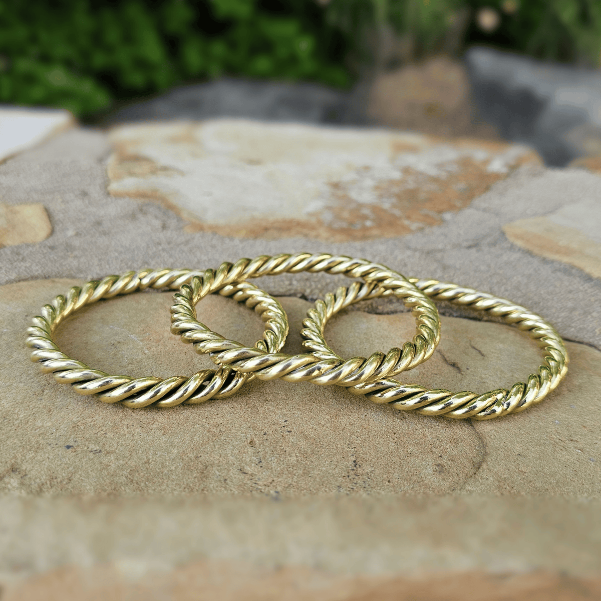 Three gold rope-style bangles stacked together on a natural stone surface with a blurred outdoor background. Shop Kamawe