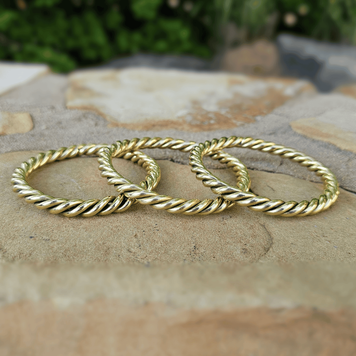 Three gold-toned twisted rope bangles displayed together on a natural stone surface with a blurred green outdoor background. Shop Kamawe