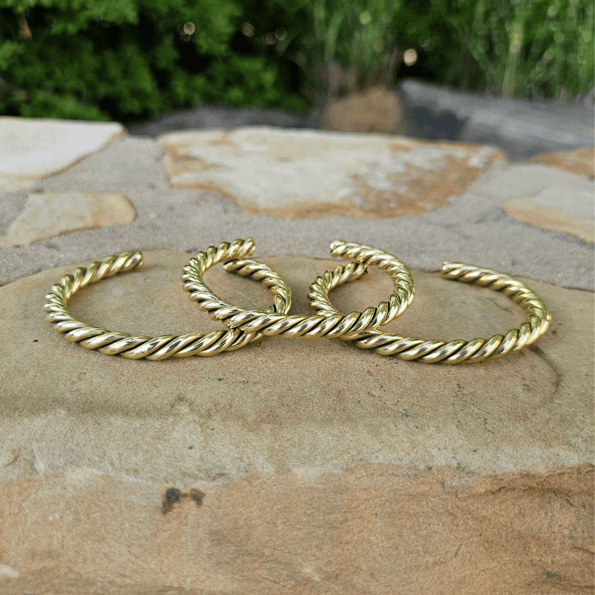 Three twisted rope-style gold bangles or bracelets arranged on a stone surface outdoors, with blurred greenery in the background. Shop Kamawe
