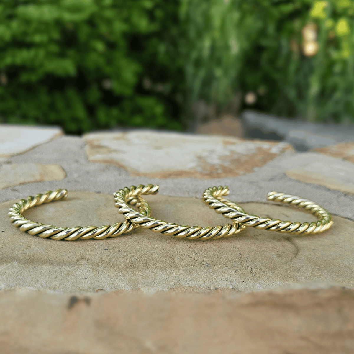 Three gold-toned twisted rope cuff earrings resting on a stone surface with a blurred green garden background. Shop Kamawe