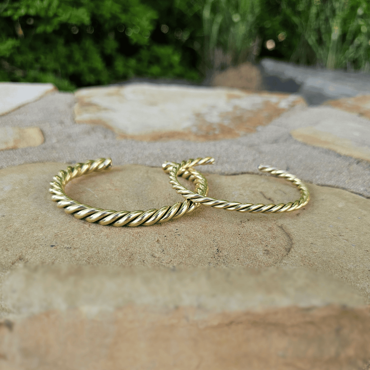 Two gold twisted rope cuff bracelets resting on a stone surface with a blurred green garden background. Shop Kamawe