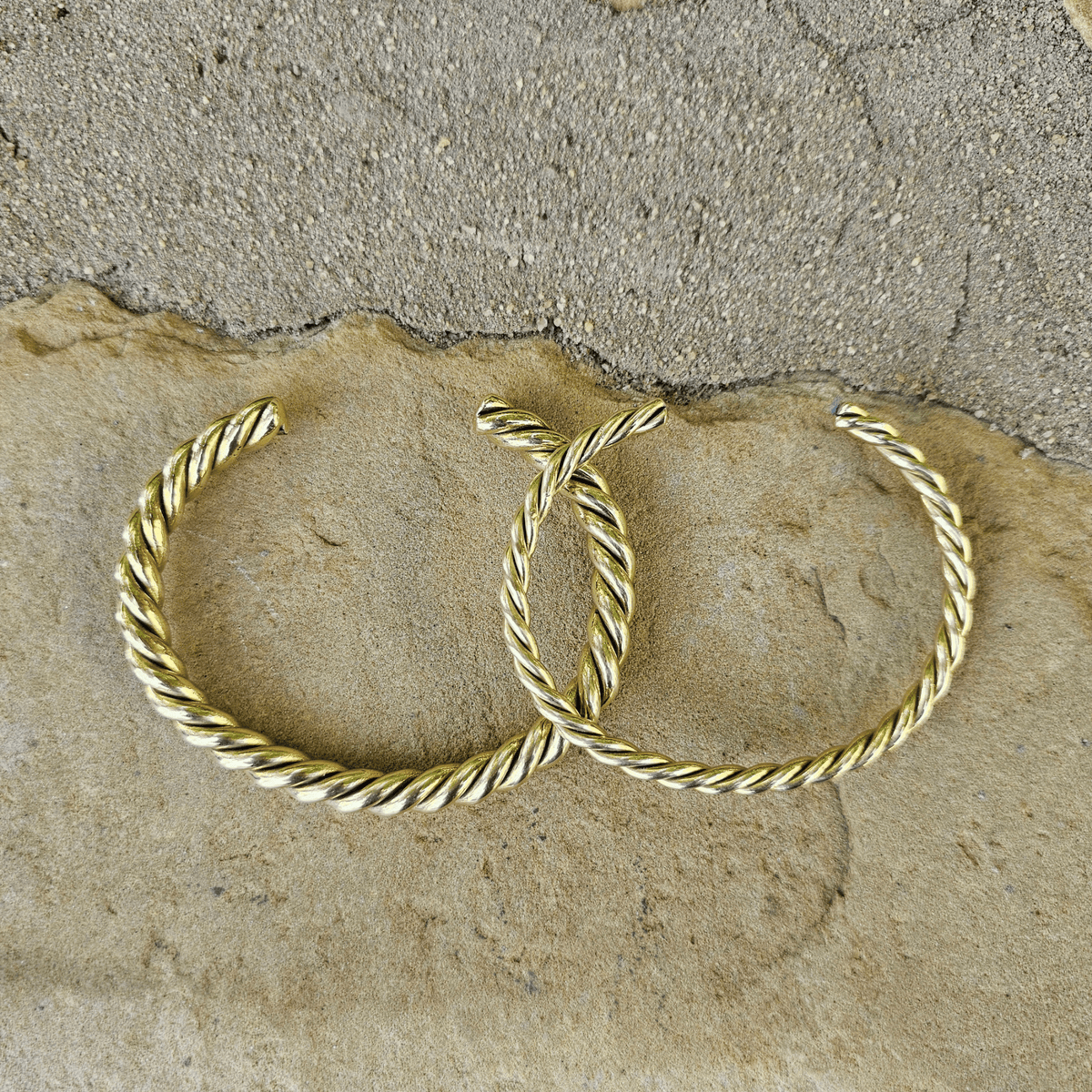 A pair of gold-toned twisted hoop earrings resting on a textured beige stone surface. Shop Kamawe