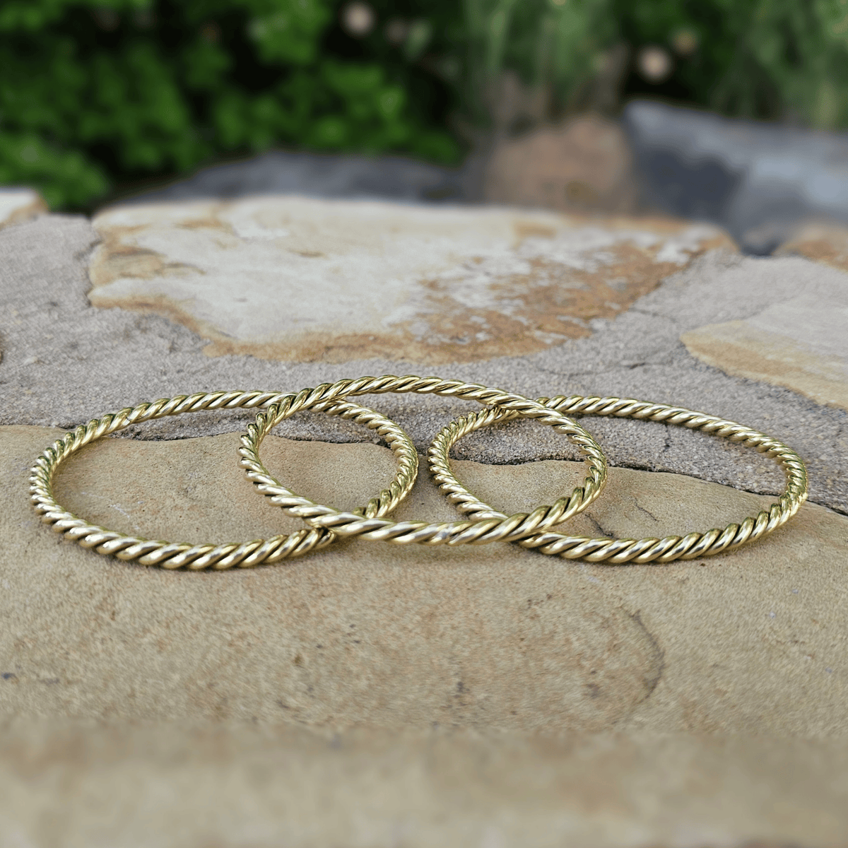 Three gold-toned twisted rope bangles arranged on a natural stone surface with a blurred outdoor background. Shop Kamawe