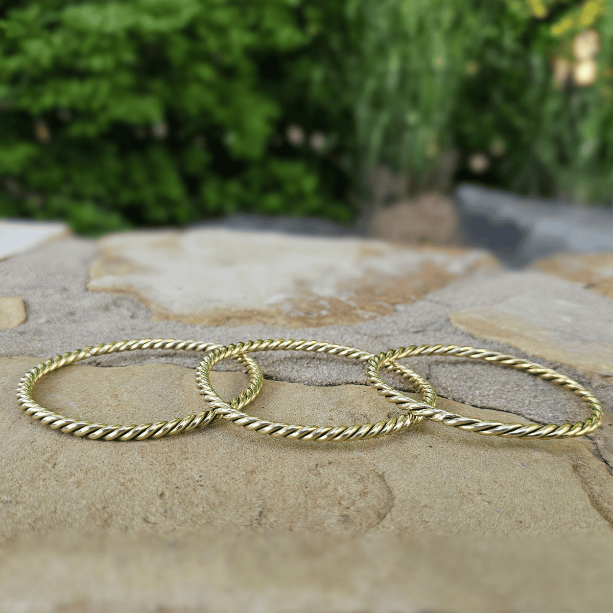 Three thin gold rope-style rings lying on a stone surface with a blurred green garden background. Shop Kamawe