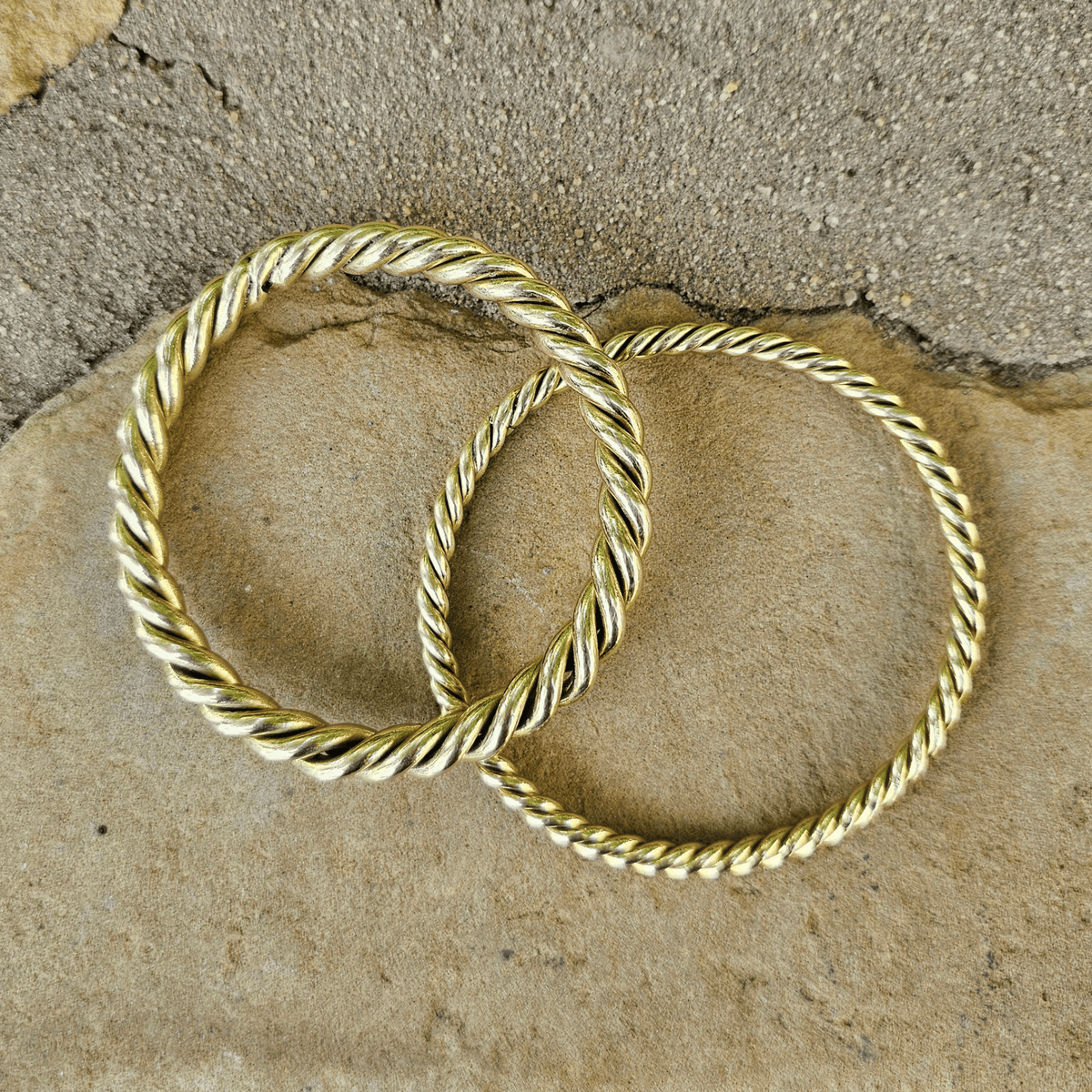 Two gold-toned twisted rope bangles resting on a textured beige stone surface. Shop Kamawe
