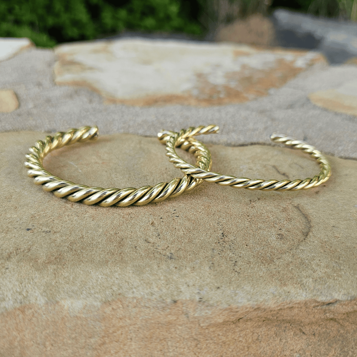Two gold-toned twisted cable hoop earrings resting on a textured stone surface with a blurred outdoor background. Shop Kamawe