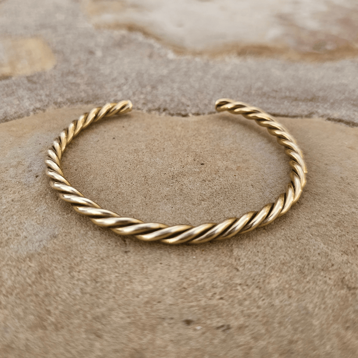 A twisted gold-tone metal cuff bracelet, likely made of copper or gold, displayed on a textured tan stone surface. Shop Kamawe