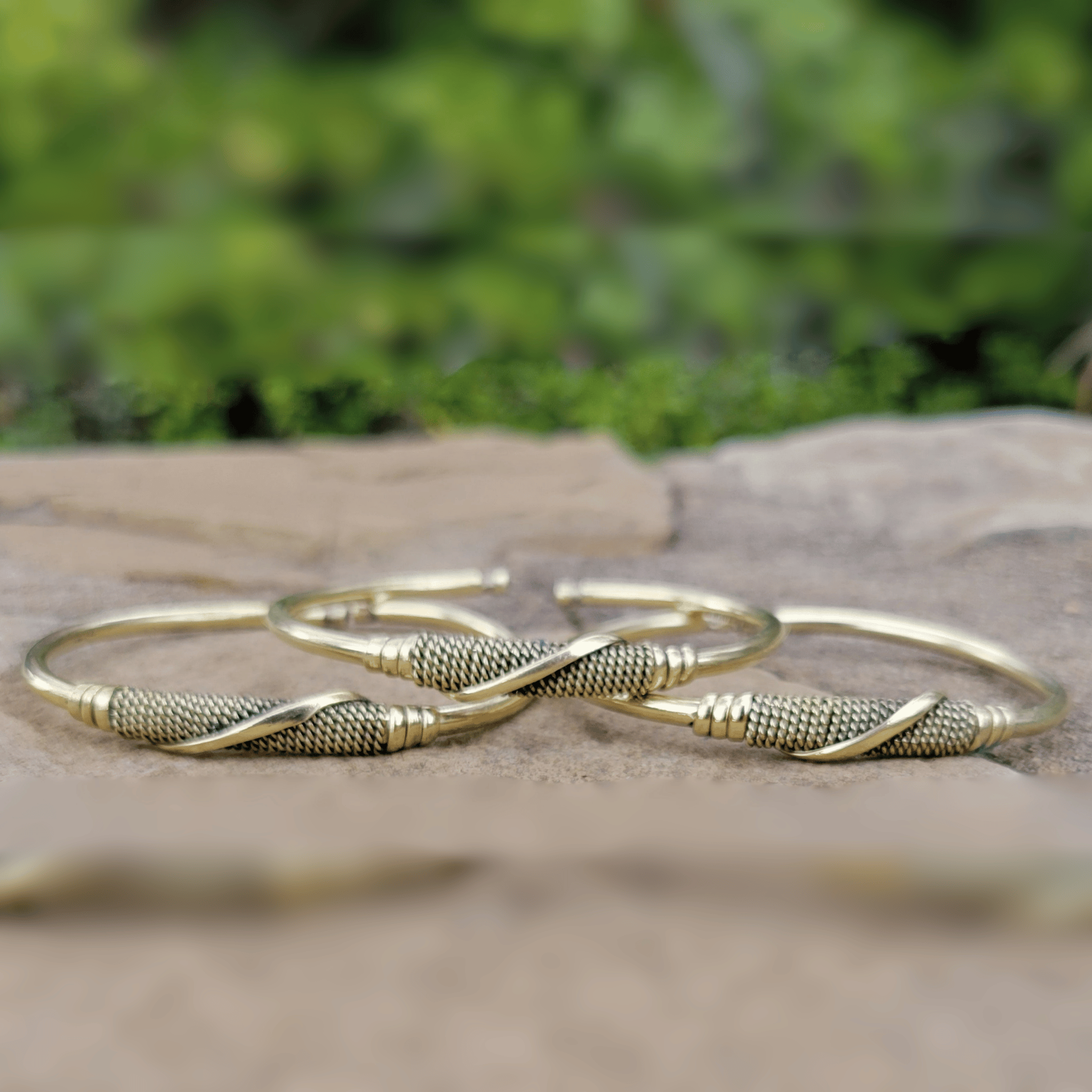 Three gold-toned metal bangles with textured mesh centers and twisted wrap details, arranged on a stone surface with a blurred green outdoor background. Shop Kamawe