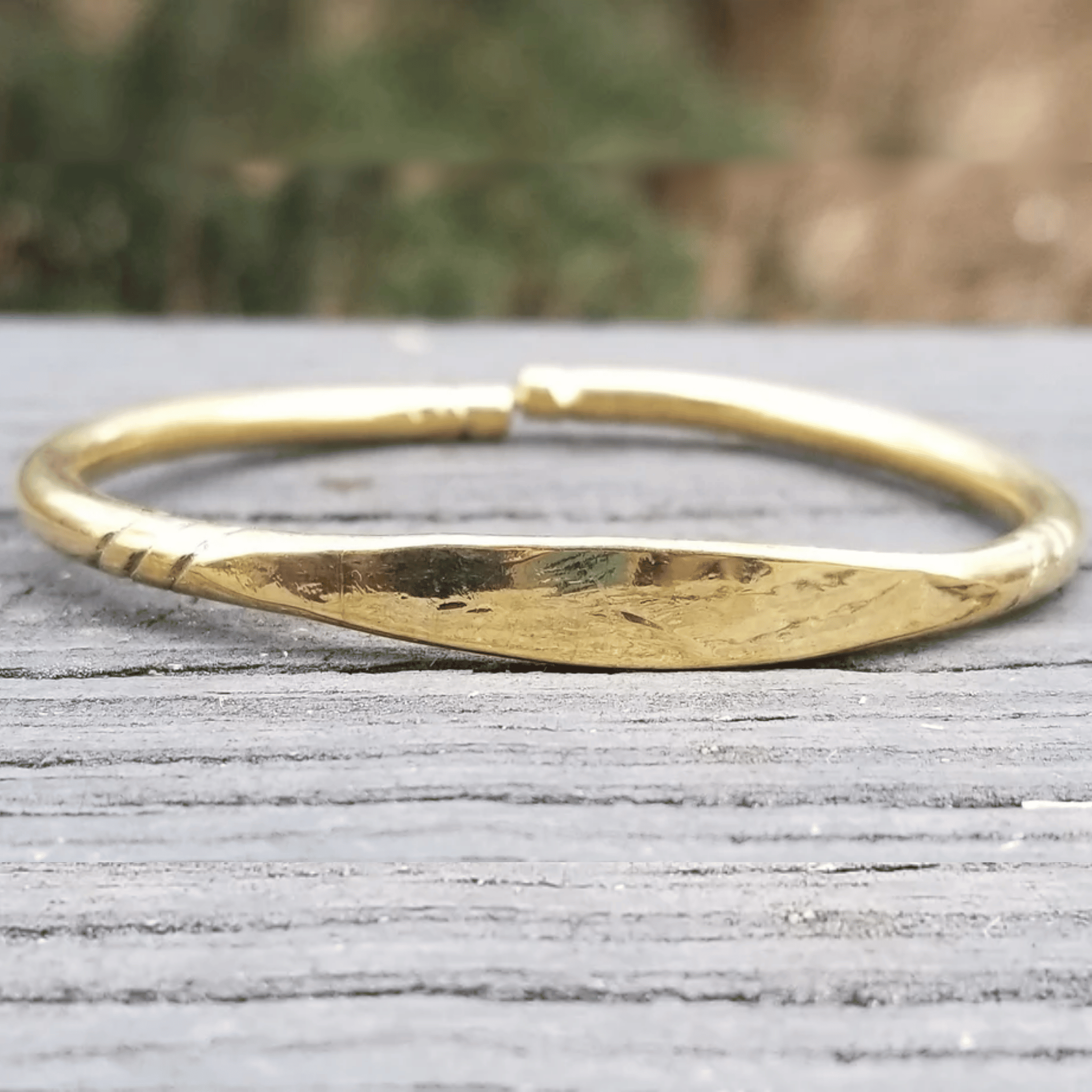 A hammered gold bangle bracelet resting on a weathered gray wooden surface with a blurred outdoor background. Shop Kamawe