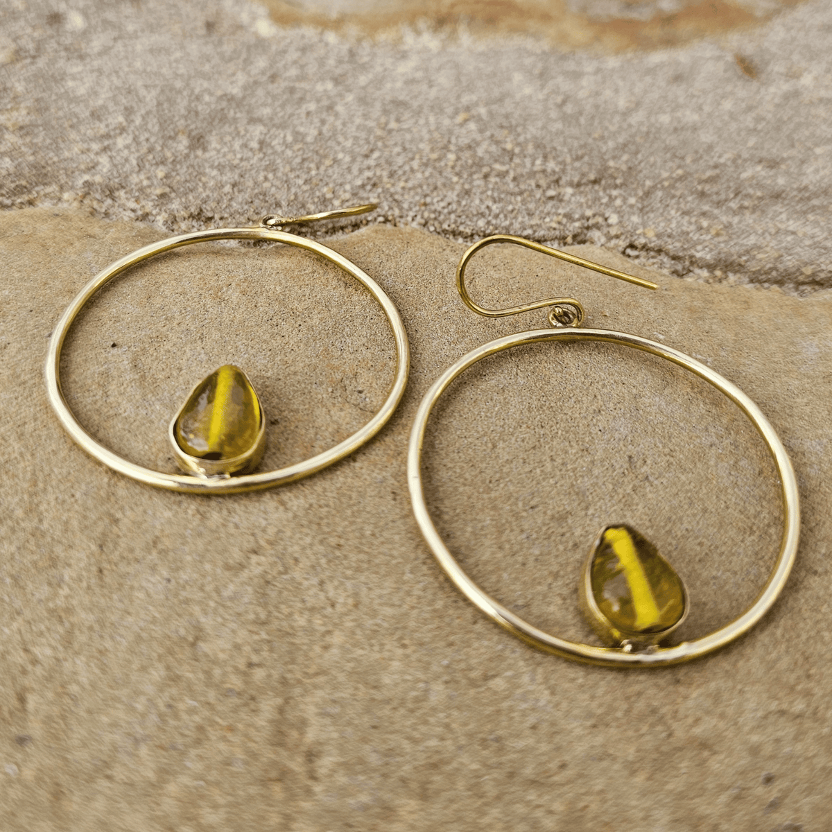 A pair of gold-tone hoop earrings with a small teardrop-shaped yellow-green bead or stone set inside each hoop, displayed on a textured beige stone surface. Shop Kamawe