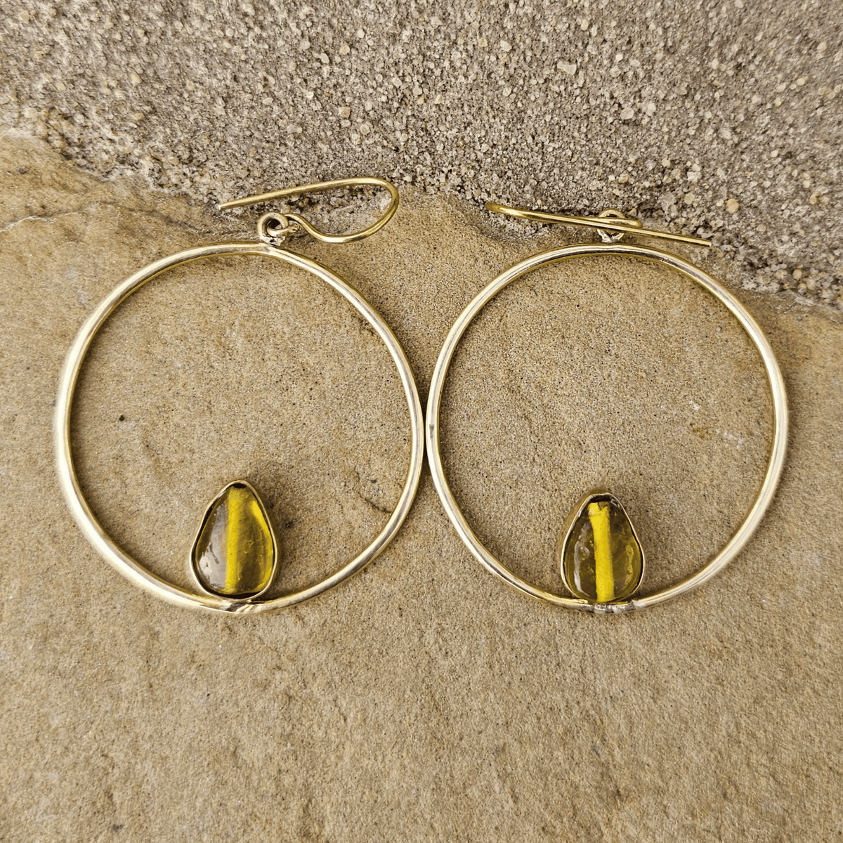 A pair of gold-tone hoop earrings with a small teardrop-shaped yellow-green bead or stone set inside each hoop, displayed on a textured beige stone surface. Shop Kamawe